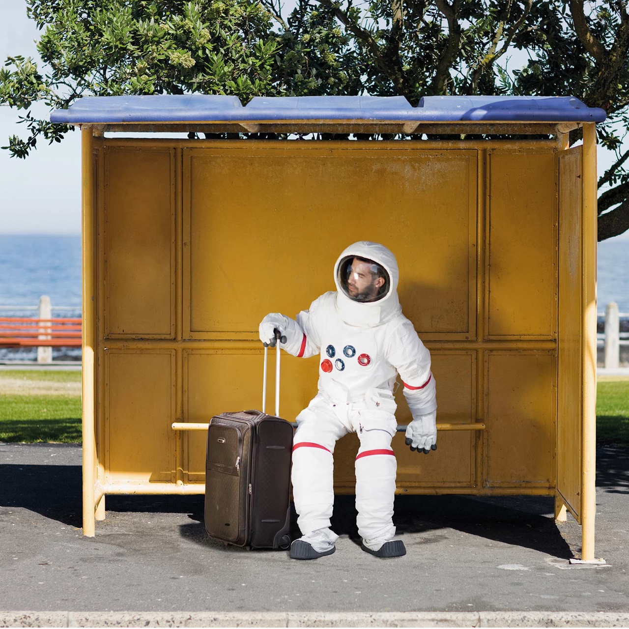 Behelmter Astronaut sitzt mit einem Rollkoffer im Bushäuschen