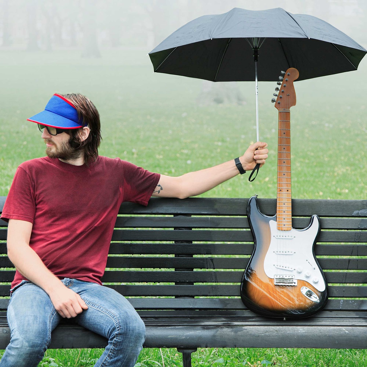 Junger Mann mit Bart sitzend auf einer Bank in einem Park und hält der daneben stehenden E-Gitarre den Regenschirm