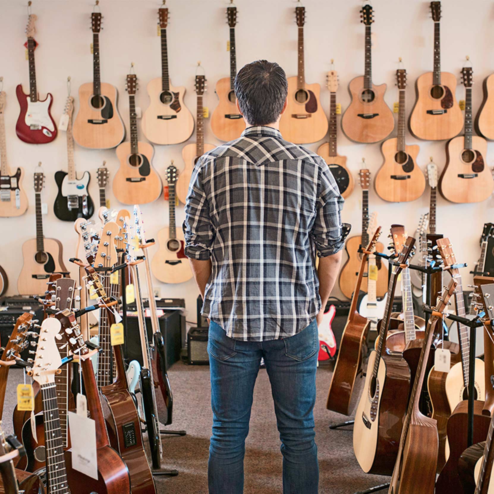 Mann steht in einem Gitarrenladen vor einer Wand voller Gitarren