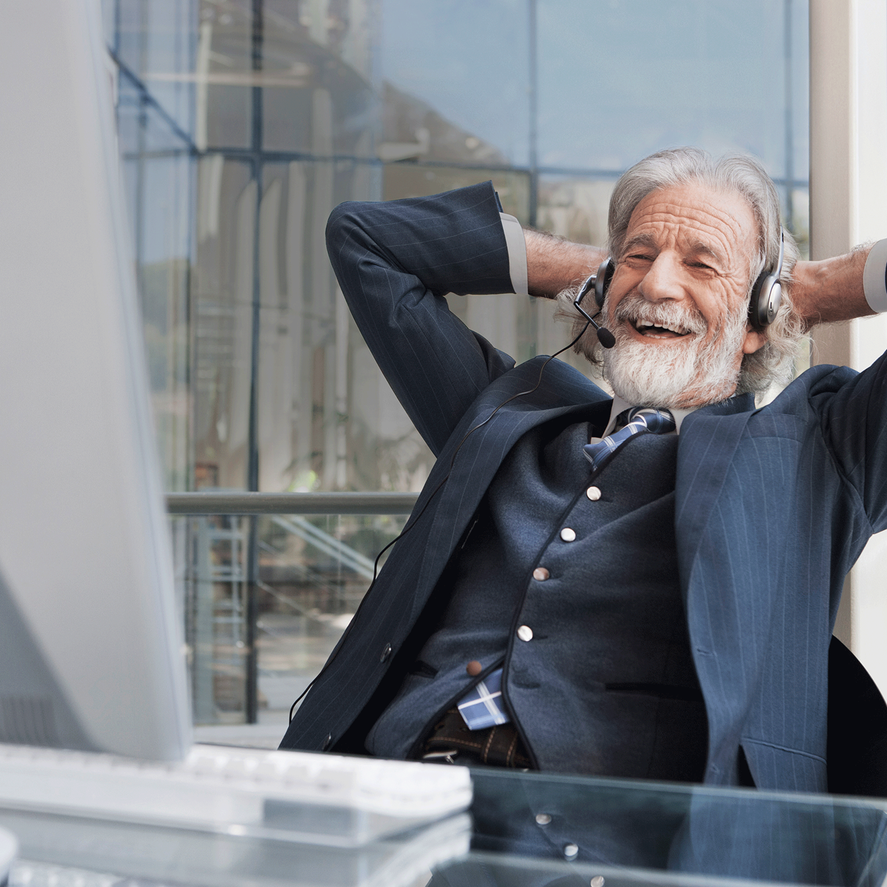 Älterer Herr im Anzug lachend sitzend vor einem Monitor und die Hände hinter dem Kopf verschränkt