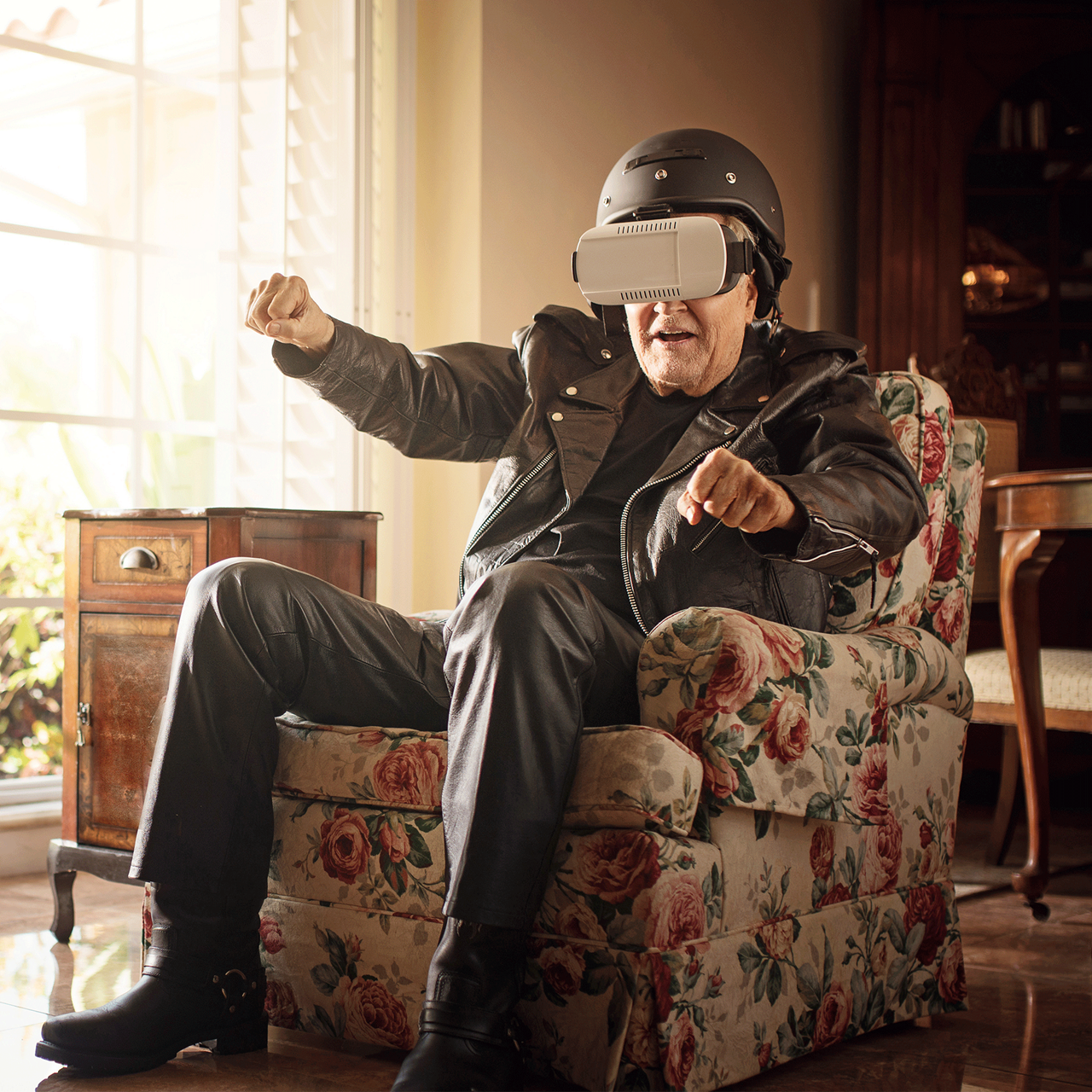 Opa in Lederkombi sitzend auf der Couch mit VR-Brille und am motorradfahren