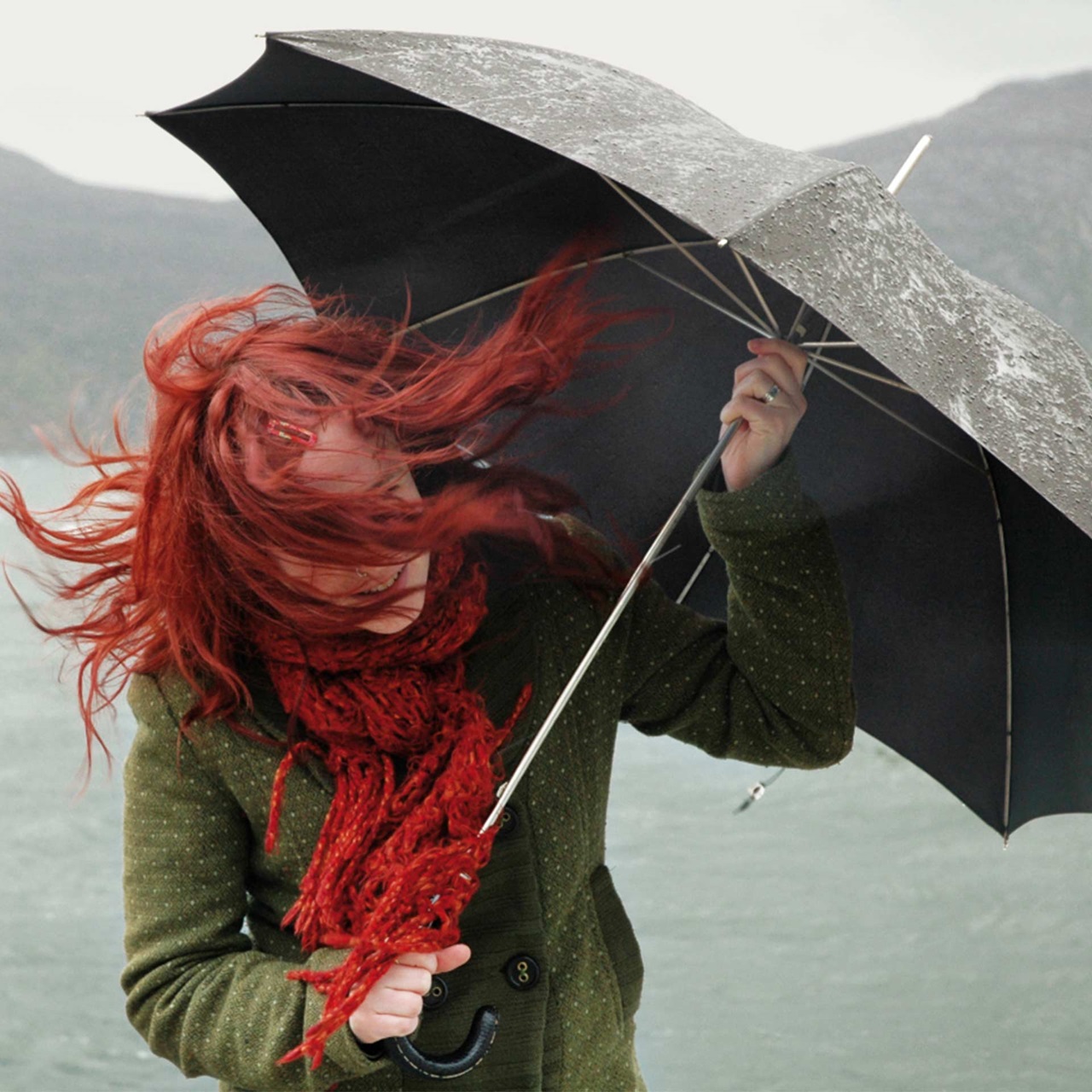 Frau mit Regenschirm steht mit flatternden Haaren im Sturm am Seeufer