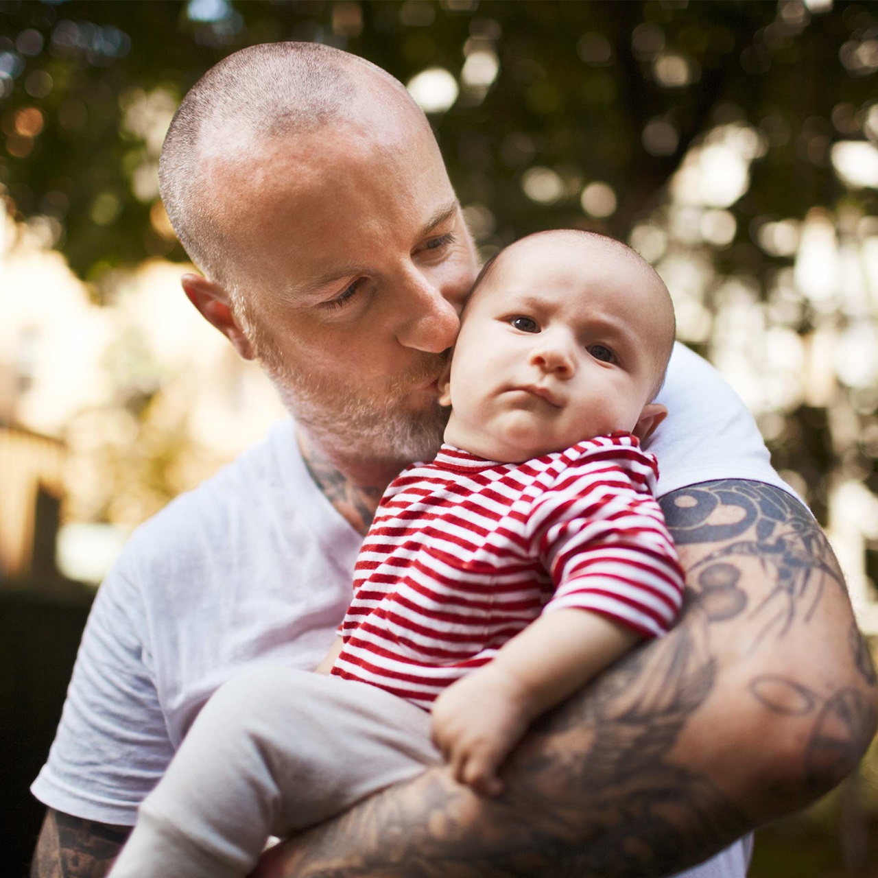 Vater mit T-Shirt und Tattoo hält seinen Babysohn und küsst ihn 