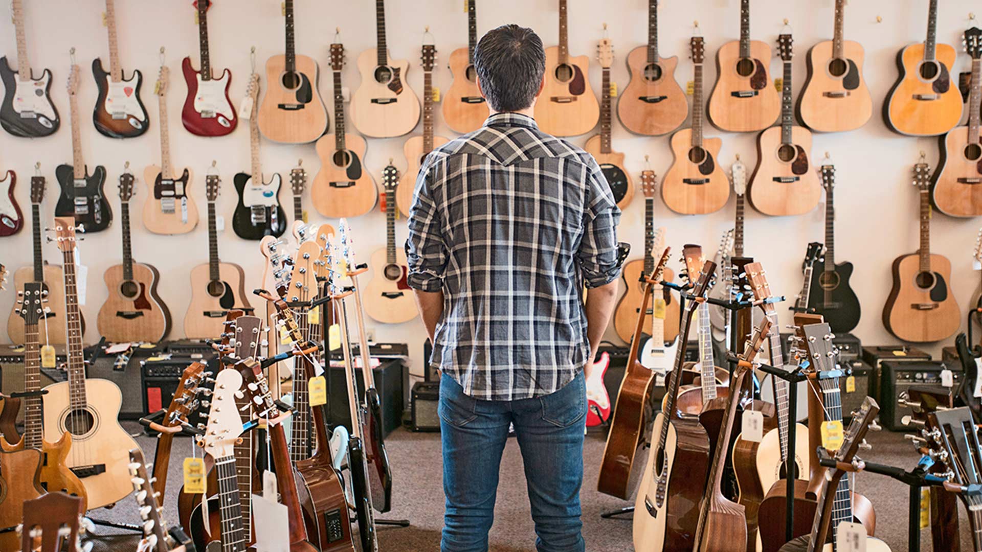 Mann steht in einem Gitarrenladen vor einer Wand voller Gitarren