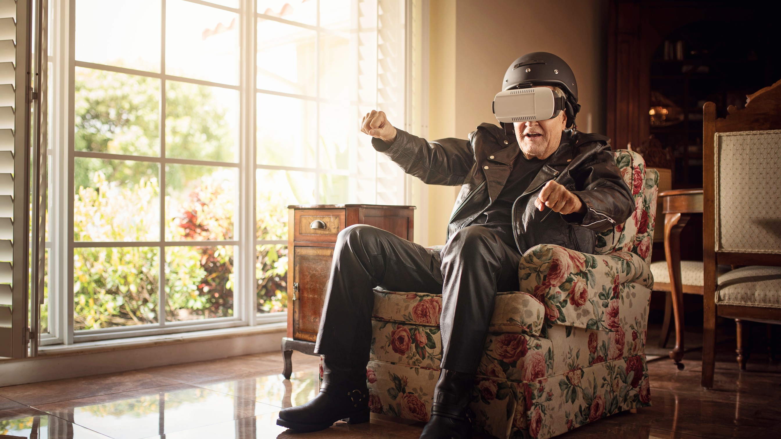 Opa in Lederkombi sitzend auf der Couch mit VR-Brille und am motorradfahren