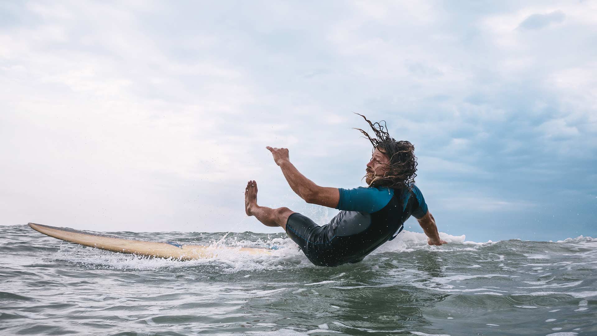Mann in Neoprenanzug und langen Haaren stürzt beim Wellenreiten vom Surfbrett