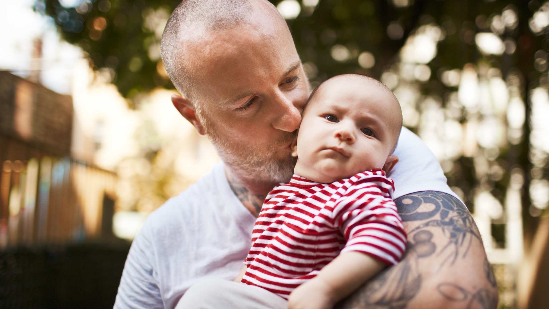 Vater mit T-Shirt und Tattoo hält seinen Babysohn und küsst ihn 