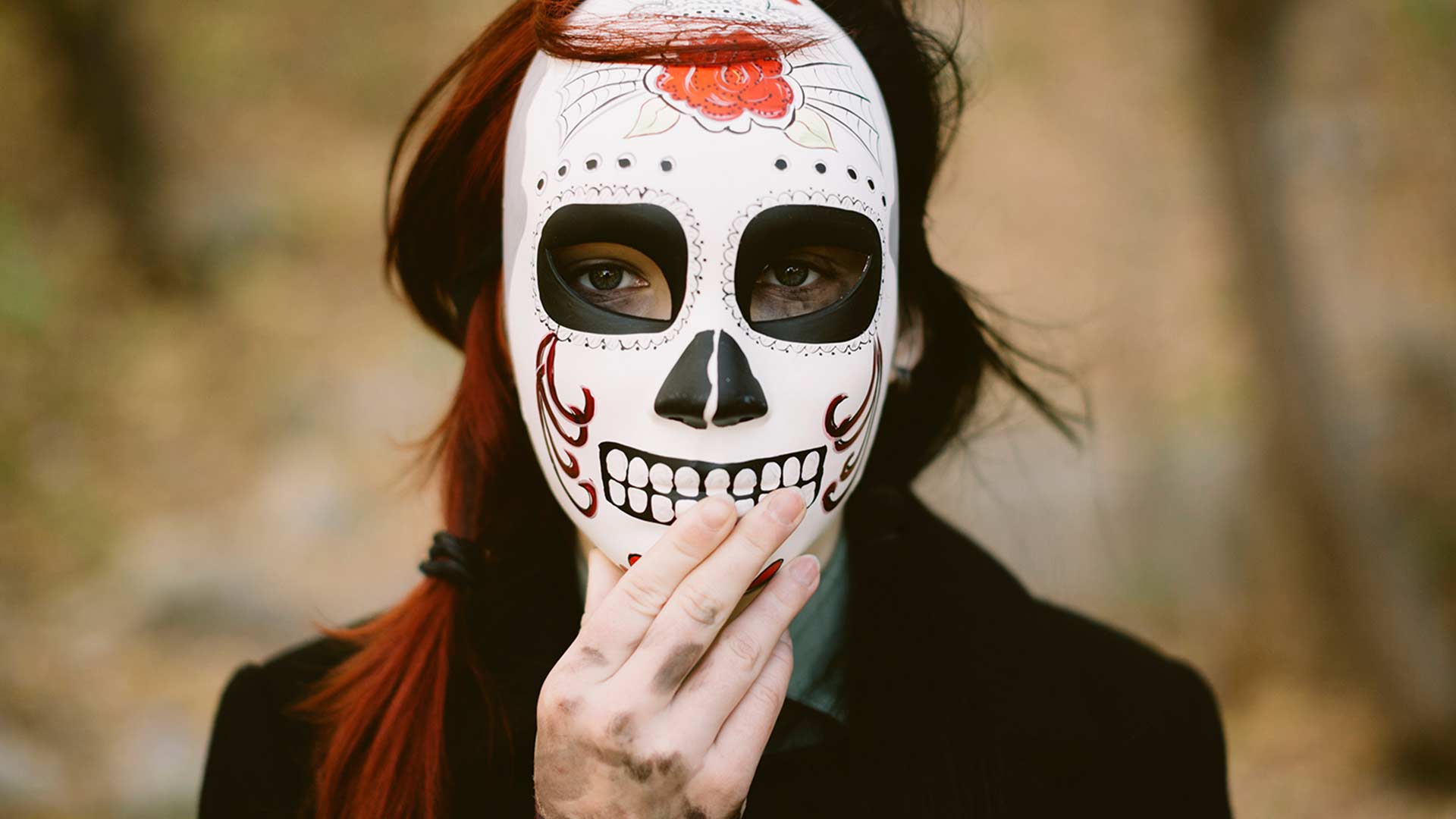 Frau mit langen Haaren hält eine mexikanische Totenmaske vors Gesicht 