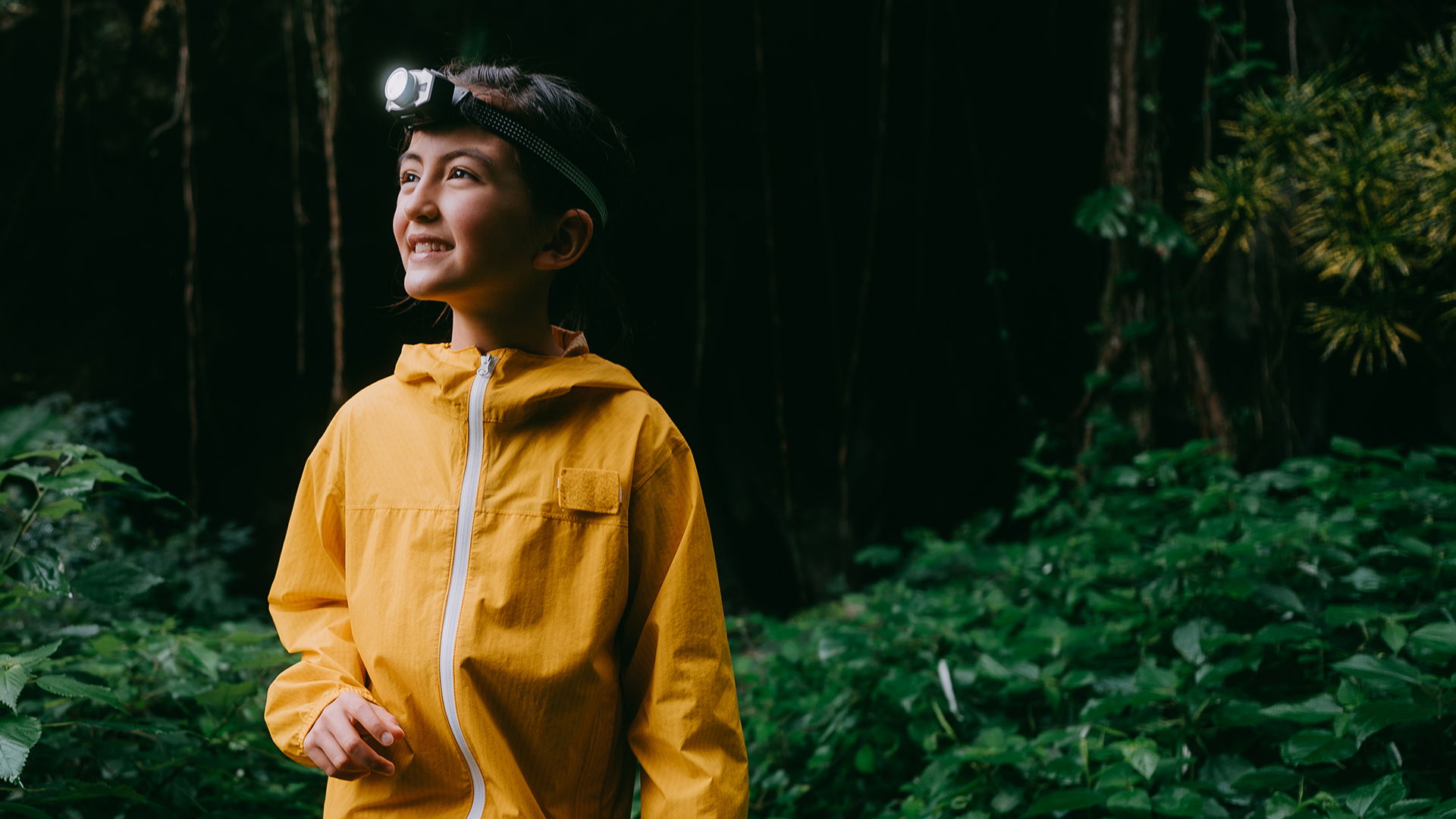 Ein Mädchen steht mit einer gelben Regenjacke im Wald und hat eine Taschenlampe auf dem Kopf und schaut zufrieden in den Wald rein.
