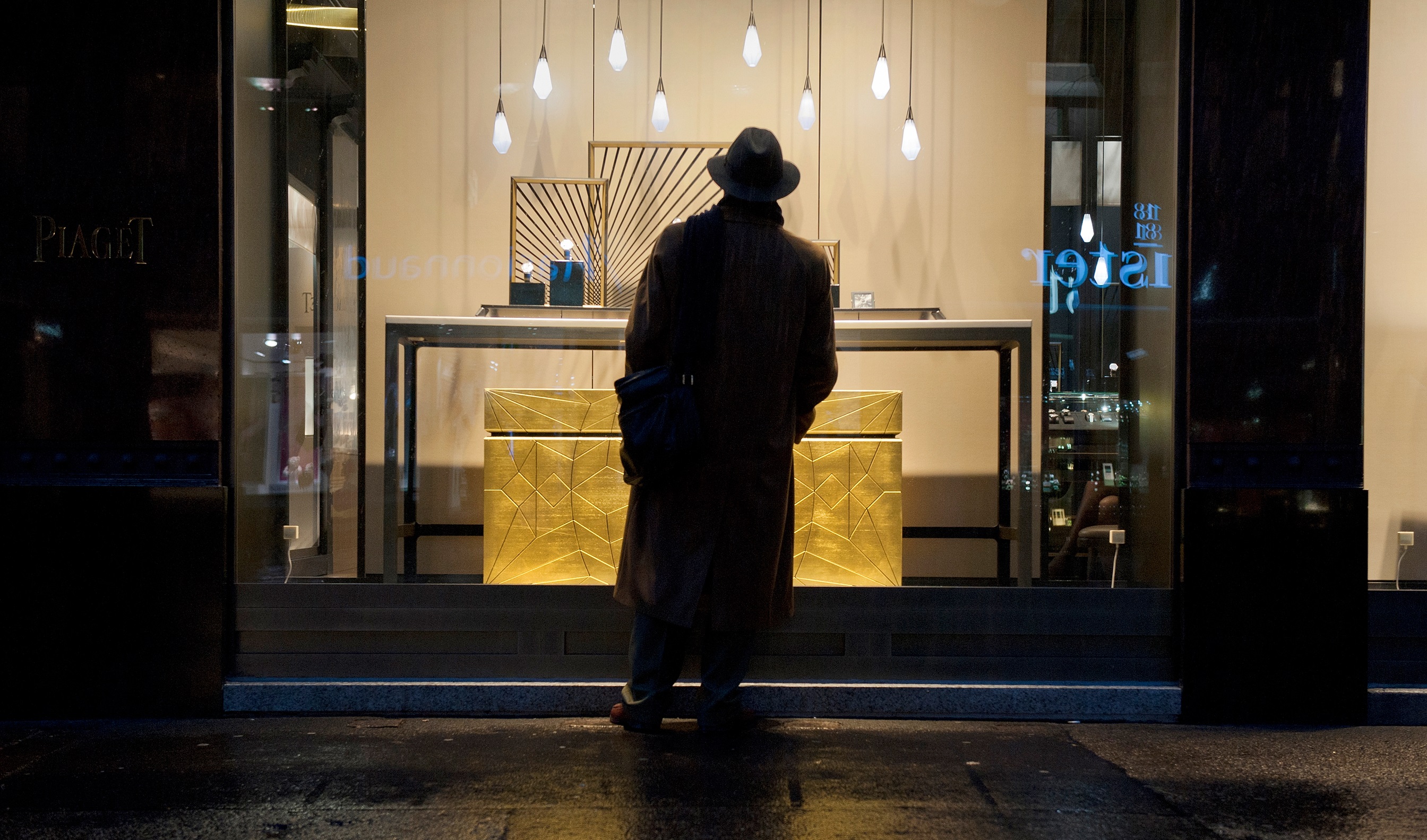 Mann mit Hut und Trenchcoat steht vor einem beleuchteten Uhrengeschäft