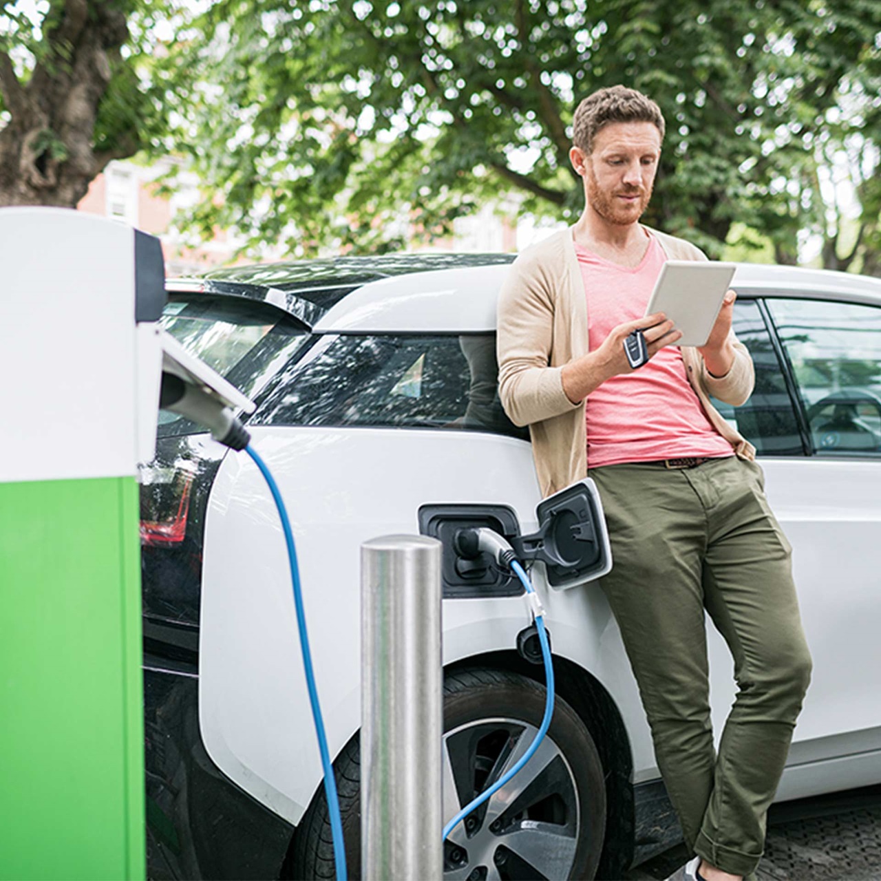 Mann mit Tablet lehnt wartend an seinem Elektroauto an der Ladestation