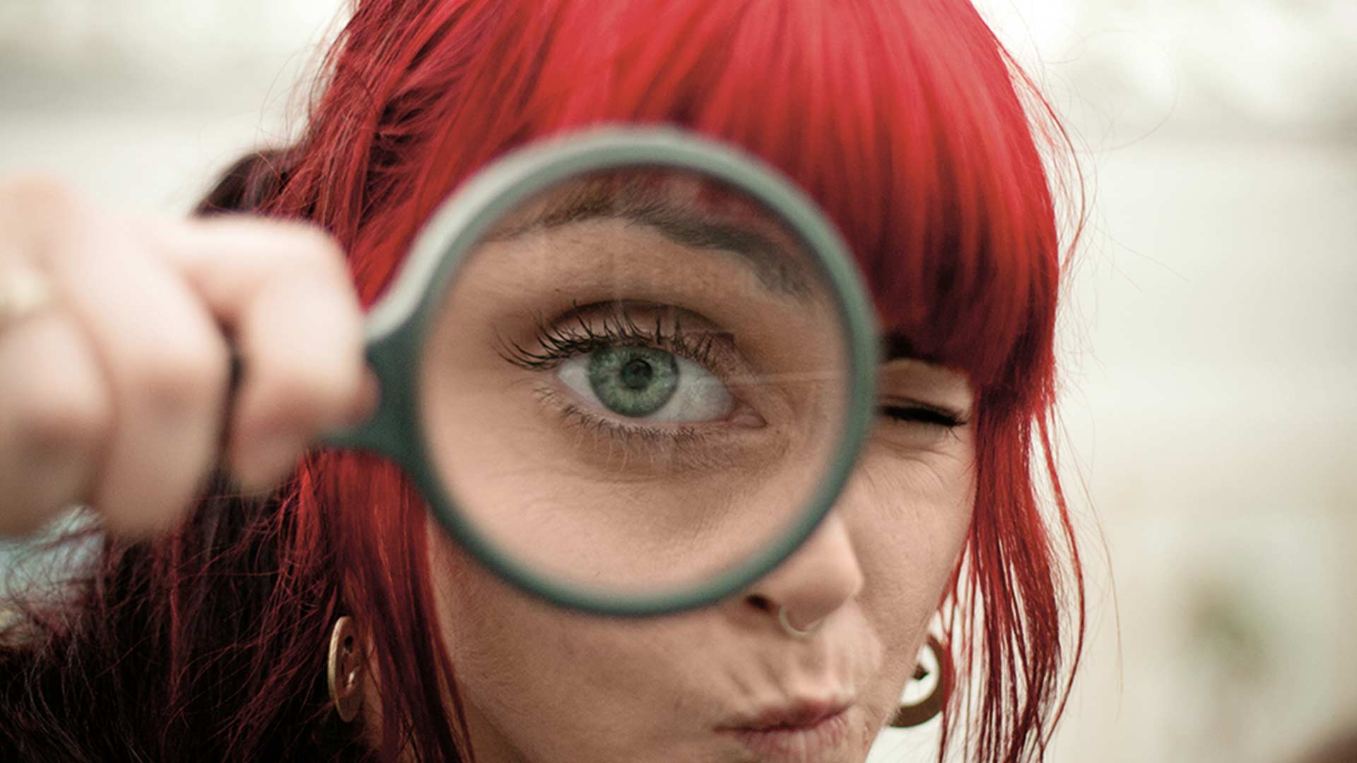 Junge Frau hält sich eine Lupe vors Auge welches riesig erscheint
