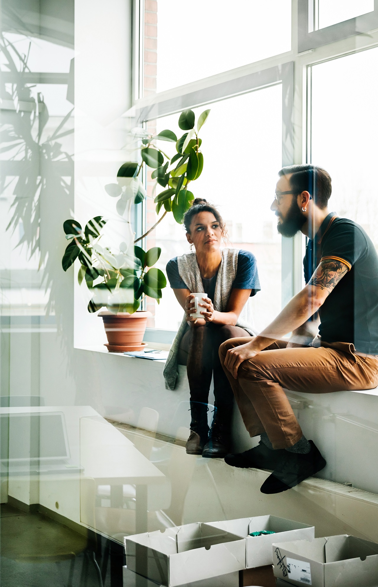 Eine Frau hält eine Tasse und sitzt neben einem jungen, tätowierten Mann in einem Büro.