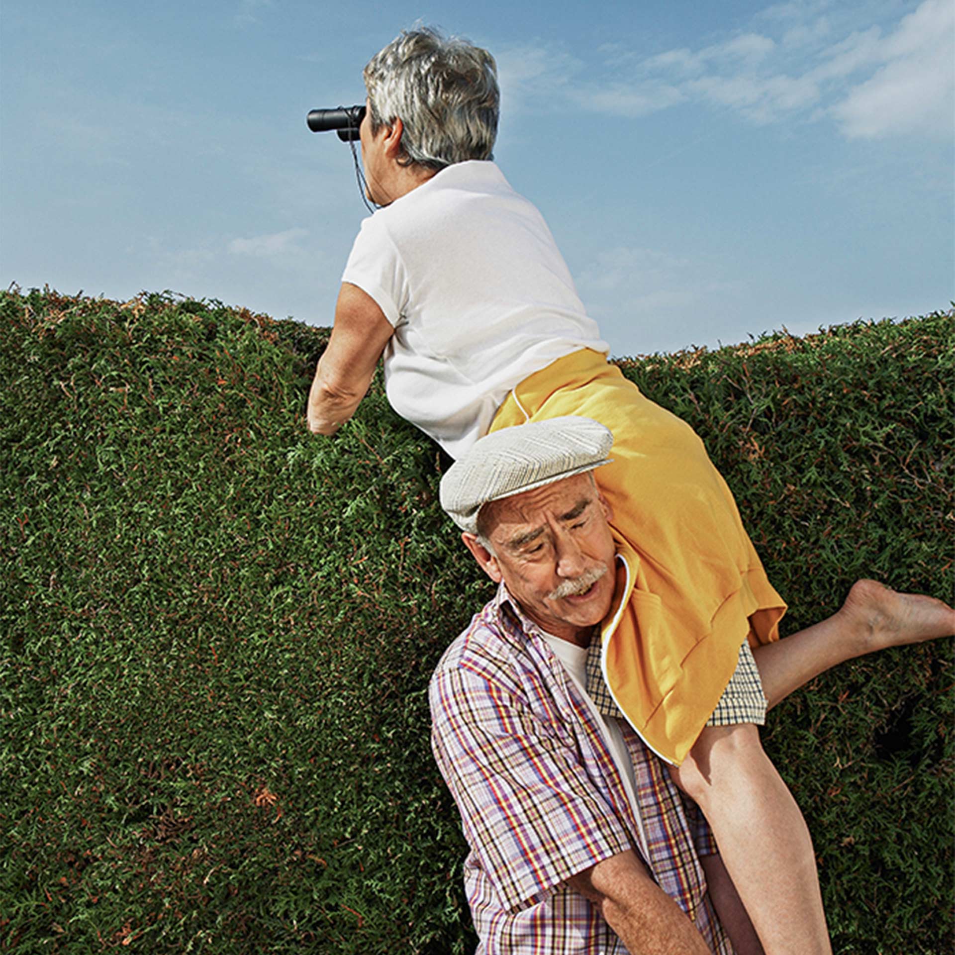 Alter Mann stemmt seine Frau hoch welche mit dem Feldstecher über Hecke schaut