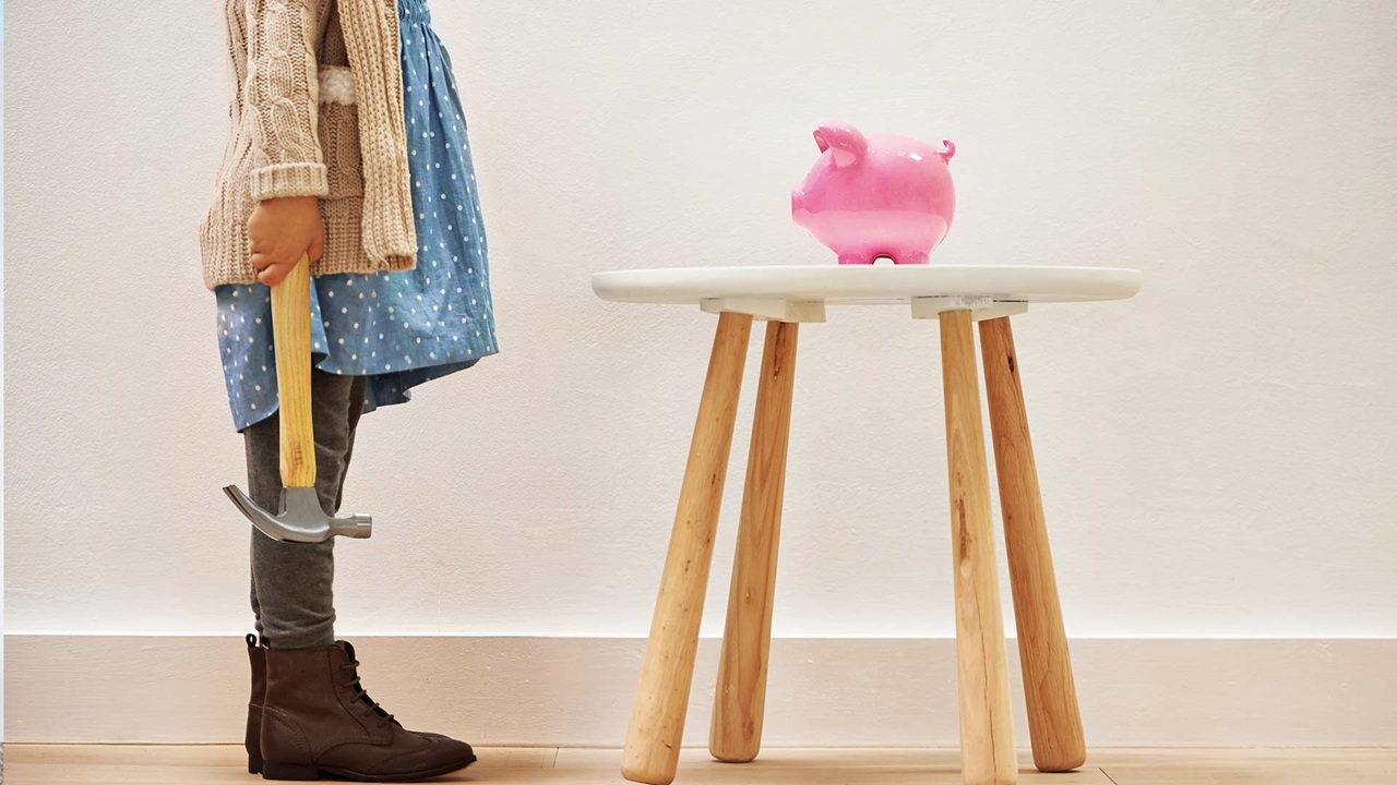 Mädchen mit Hammer steht vor einem Tischchen mit einem pinken Sparschwein